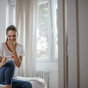 Mujer sentada en la cama sonriendo y mirando un test de embarazo