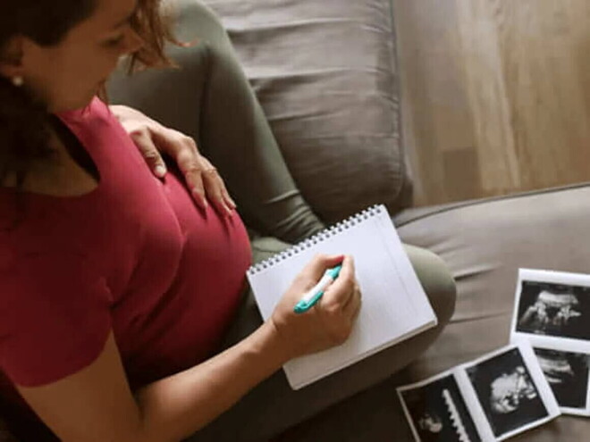 Mujer embarazada de sentada en un sofá, anotando nombres para bebés varones en un cuaderno con imágenes de ecografía a su lado.