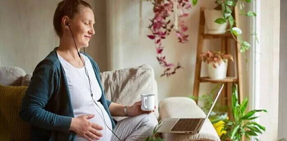 Mujer embarazada sentada en el sofá viendo una película con su laptop y audífonos para acelerar la dilatación.